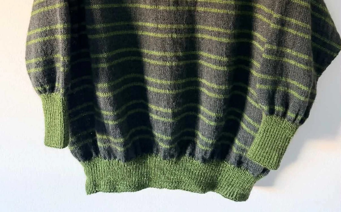 The bottom of a knitted, not yet blocked Collioure sweater, very dark green with fine bright green stripes, with its balloon sleeves gathered into ribbed cuffs and its gathered hem